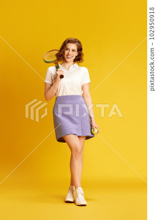 Full-length portrait of young, smiling, beautiful girl in sportswear posing with tennis racket against yellow studio background 102950918