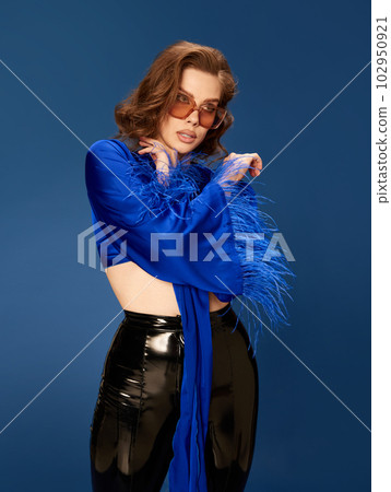 Portrait of attractive young brunette woman in trendy sunglasses, stylish top and leather pants posing against blue studio background 102950921