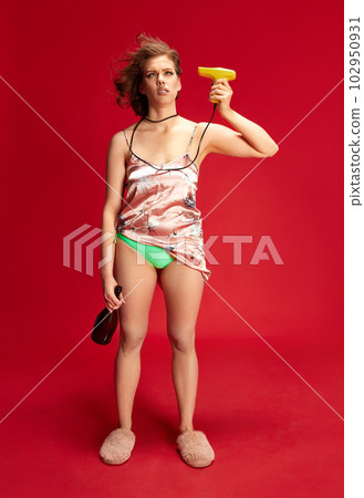 Portrait of young beautiful woman with emotionless face, blowing with hairdryer, holding wine bottle against red studio background Portrait of young beautiful woman with emotionless face, blowing with hairdryer, holding wine bottle against red studio background 102950931