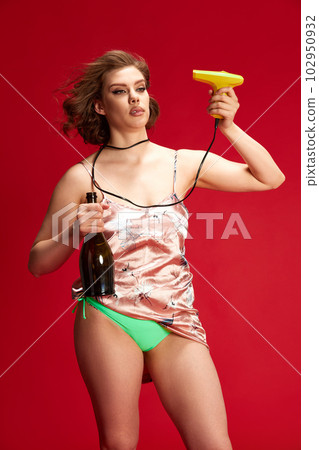 Portrait of young beautiful woman with emotionless face, blowing with hairdryer, holding wine bottle against red studio background 102950932
