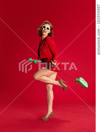 Carefree mood. Portrait of pretty young women in classical elegant suit and sunglasses, smiling, holding phone against red studio background Carefree mood. Portrait of pretty young women in classical elegant suit and sunglasses, smiling, holding phone against red studio background 102950997