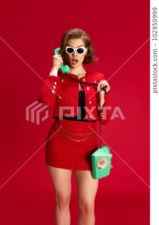 Portrait of attractive, beautiful young woman in elegant suit talking on phone with shocked, impressed face against red studio background 102950999