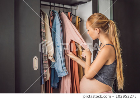 A pregnant woman has nothing to wear. A pregnant woman stands in front of a closet with clothes and does not know what to wear because the clothes do not fit on her A pregnant woman has nothing to wear. A pregnant woman stands in front of a closet with clothes and does not know what to wear because the clothes do not fit on her 102952275