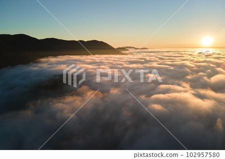 Aerial view of colorful sunrise over white dense fog with distant dark silhouettes of mountain hills on horizon Aerial view of colorful sunrise over white dense fog with distant dark silhouettes of mountain hills on horizon 102957580