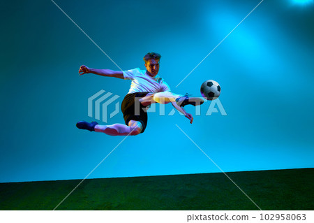 Professional man, football player in sports team uniform kicking the ball in motion over soccer field background in neon light. Scoring a goal. 102958063