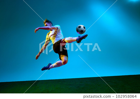 One sportsman, soccer player wearing yellow and black uniform hitting the ball in the air over football field in neon light. Concept of action, energy, sport 102958064
