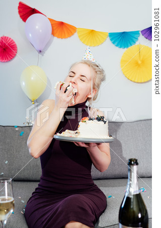 Woman in festive dress and party cap eating birthday cake with her hands, her face stained cream. A woman stuffing her face with cake. Crazy celebrating birthday party at home. Selective focus. Woman in festive dress and party cap eating birthday cake with her hands, her face stained cream. A woman stuffing her face with cake. Crazy celebrating birthday party at home. Selective focus. 102961081