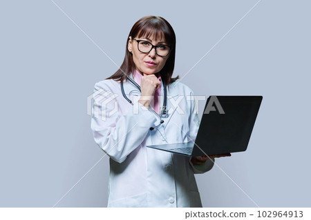 Mature female doctor using laptop on grey background Mature female doctor using laptop on grey background 102964913