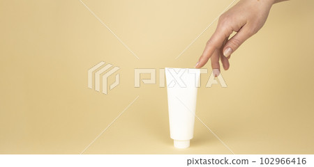 Layout for branding cosmetics. A tender female hand touches a tube of skin care cream on a beige background. Layout for branding cosmetics. A tender female hand touches a tube of skin care cream on a beige background. 102966416