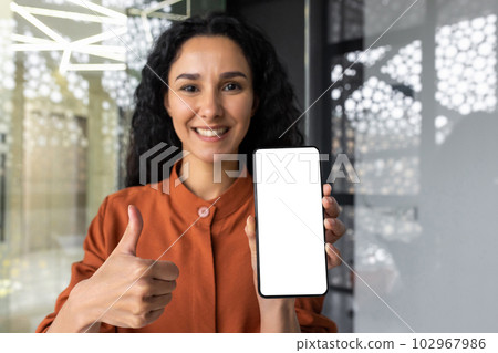 Successful Latin American businesswoman at workplace inside office sitting at table showing smartphone screen to camera, joyfully recommending app with thumbs up. 102967986