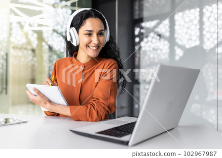 Latin american business woman with curly hair and headphones watching online training course at workplace, woman writing information happy and satisfied with the results of professional development. 102967987
