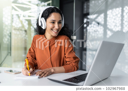 Latin american business woman with curly hair and headphones watching online training course at workplace, woman writing information happy and satisfied with the results of professional development. 102967988