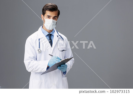 A male doctor in a white coat and a medical mask with a notepad for notes and a patient card in his hand looks at the camera on a gray isolated background, copy space, space for text A male doctor in a white coat and a medical mask with a notepad for notes and a patient card in his hand looks at the camera on a gray isolated background, copy space, space for text 102973315