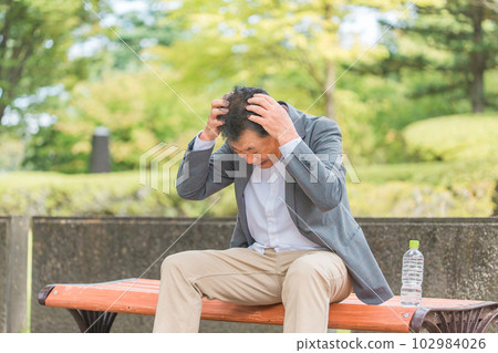 Elderly Asian man holding his head in distress on a park bench 102984026