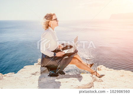 Digital nomad, Business woman working on laptop by the sea. Pretty lady typing on computer by the sea at sunset, makes a business transaction online from a distance. Freelance, remote work on vacation 102986560