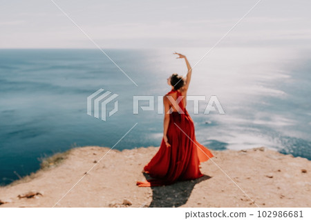 Woman in red dress on sea. Side view a Young beautiful sensual woman in a red long dress posing on a rock high above the sea on sunset. Girl on the nature on blue sky background. Fashion photo. Woman in red dress on sea. Side view a Young beautiful sensual woman in a red long dress posing on a rock high above the sea on sunset. Girl on the nature on blue sky background. Fashion photo. 102986681