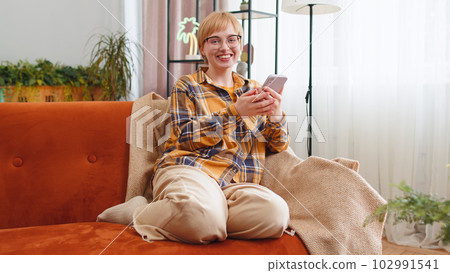 Young woman sitting on sofa at home, using smartphone share messages on social media application 102991541