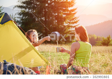 Two women travellers camping outdoors. Young women sitting in campsite, having break. Friends sitting on grass, talking, smiling, pouring water, making tea. Concept of tourism and hiking in mountains. 102991601