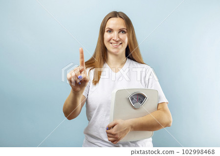 Woman holding white scale on blue background showing number one with finger Woman holding white scale on blue background showing number one with finger 102998468