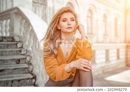 A middle-aged woman looks like a good blonde with curly beautiful hair and makeup on the background of the building. She is wearing a yellow sweater. A middle-aged woman looks like a good blonde with curly beautiful hair and makeup on the background of the building. She is wearing a yellow sweater. 102999270