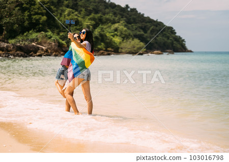 Couple of cheerful women with rainbow flag standing on beach Couple of cheerful women with rainbow flag standing on beach 103016798
