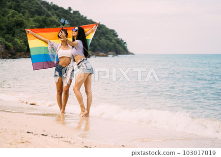 Couple of cheerful women with rainbow flag standing on beach Couple of cheerful women with rainbow flag standing on beach 103017019