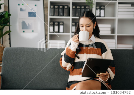 Beautiful asian woman holding and using digital tablet while drinking coffee at home office and cafe Beautiful asian woman holding and using digital tablet while drinking coffee at home office and cafe 103034792