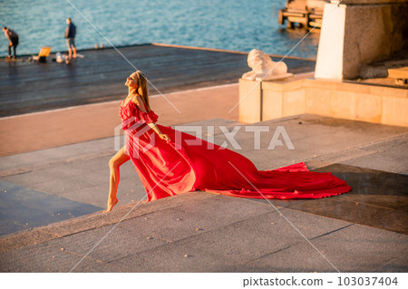 Sunrise red dress. A woman in a long red dress against the backdrop of sunrise, bright golden light of the sun's rays. The concept of femininity, harmony. 103037404