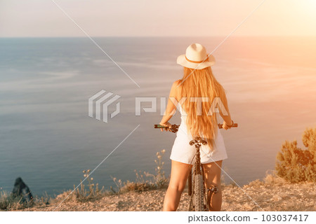 Woman travel bike sea. Happy woman cyclist sitting on her bike, enjoying the beautiful mountain and sea landscape, signifying the idea of an adventurous bike ride. 103037417