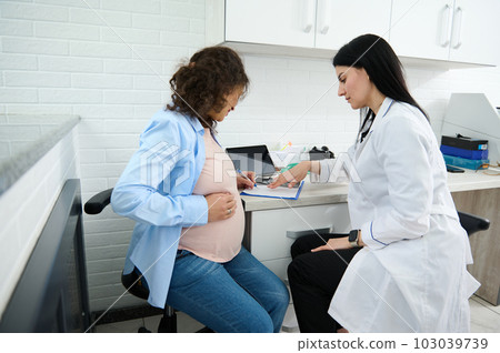 Young pregnant woman putting hand on her belly, sitting in a doctor's office and signing a contract with a medical clinic for pregnancy management. Obstetrics and gynecology. Medical insurance concept 103039739