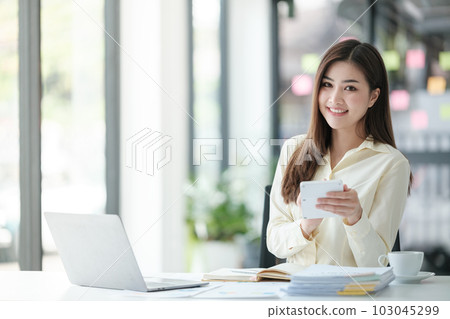 Photo of attractive woman sitting at desk working on computer looking at screen, smiling, wearing yellow shirt and glasses, typing on keyboard, holding a cup of coffee. 103045299