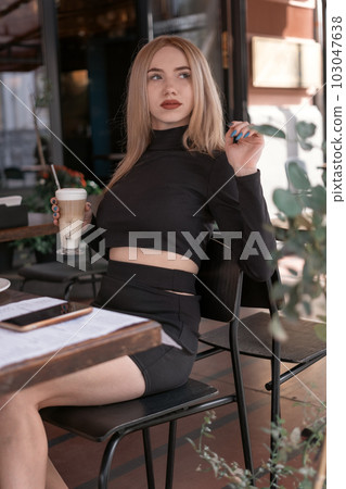 Stylish young woman in tight black clothes sits at table in cafe and holds latte or cappuccino coffee. Vertical frame. Stylish young woman in tight black clothes sits at table in cafe and holds latte or cappuccino coffee. Vertical frame. 103047638