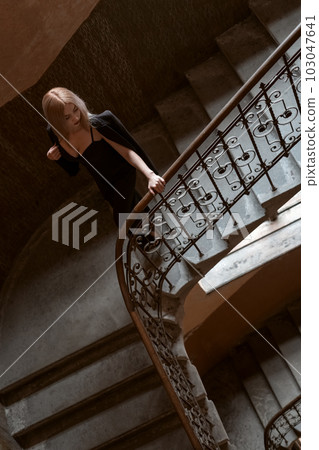 Attractive young woman with deep neckline throws jacket over her shoulders and goes down an old staircase. Top view. Vertical frame. 103047641