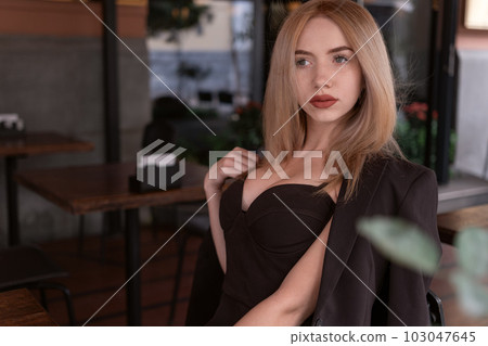 Stylish young woman in black outfit with deep neckline sits in cafe. Sexy fair-haired model in the cafeteria. Stylish young woman in black outfit with deep neckline sits in cafe. Sexy fair-haired model in the cafeteria. 103047645
