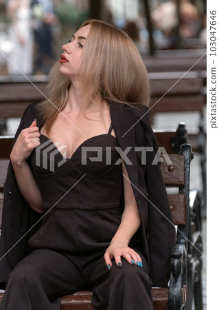 Sexy young attractive blonde woman in black suit with deep neckline on bench. Vertical frame 103047646