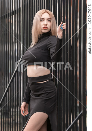 Stylish young woman in short black top and bare stomach. Blonde sexy girl poses near metal fence. Vertical frame. 103047648