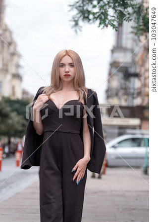 Elegant lady in business suit walks around the city. Young woman is blonde in black suit with deep neckline. Vertical frame. 103047649