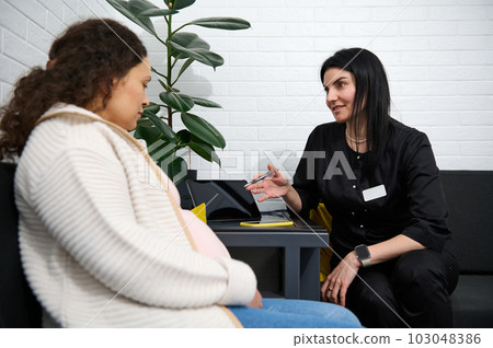 Adult pregnant female patient talking with doctor about test result diagnosis and ultrasound scan of baby at hospital corridor, listening to doctor's prescription. Obstetrics and gynecology concept Adult pregnant female patient talking with doctor about test result diagnosis and ultrasound scan of baby at hospital corridor, listening to doctor's prescription. Obstetrics and gynecology concept 103048386