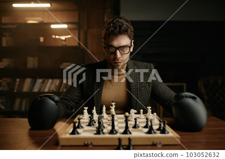 Angry young man wearing boxer gloves sitting at table with chessboard Angry young man wearing boxer gloves sitting at table with chessboard 103052632