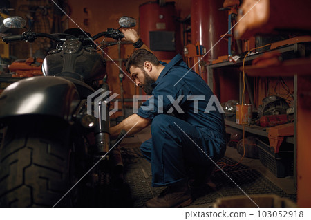 Mechanic working with motorcycle engine in workshop while sitting nearby vehicle 103052918