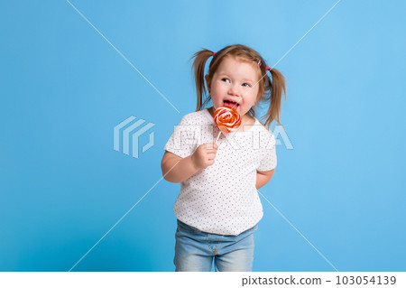 Beautiful little female child holding huge lollipop spiral candy smiling happy isolated on blue background. 103054139