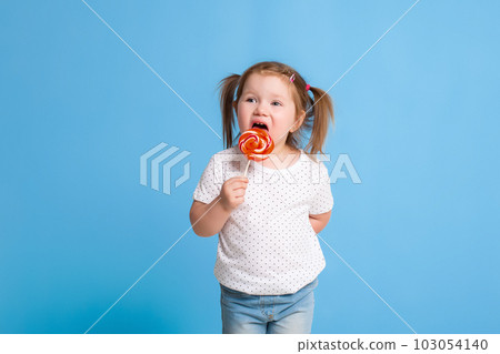 Beautiful little female child holding huge lollipop spiral candy smiling happy isolated on blue background. 103054140