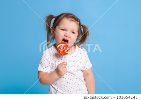 Beautiful little female child holding huge lollipop spiral candy smiling happy isolated on blue background. 103054143