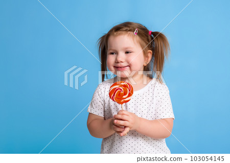 Beautiful little female child holding huge lollipop spiral candy smiling happy isolated on blue background. Beautiful little female child holding huge lollipop spiral candy smiling happy isolated on blue background. 103054145