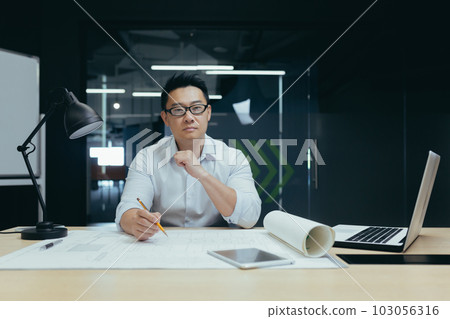 Portrait of a young Asian male designer, architect, engineer looking confidently and seriously at the camera, working in the office at a table with papers and projects. 103056316