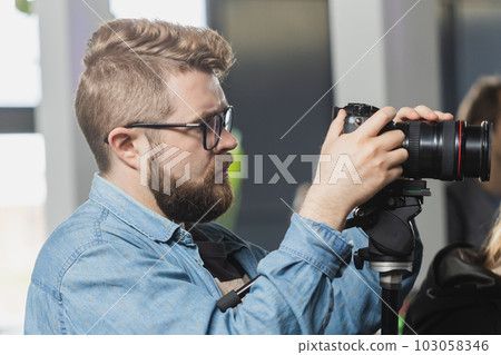 Man videomaker works on news conference modern video camera with a digital display recording an interview on event. Mass media and television and technology concept Man videomaker works on news conference modern video camera with a digital display recording an interview on event. Mass media and television and technology concept 103058346