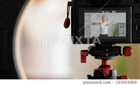 Recording online workout. Sport vlog. Cheerful woman training at home filming creating healthy lifestyle content using camera tripod. 103059969