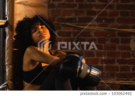 Woman fighter girl power. African american woman fighter with boxing gloves sitting on boxing ring waiting and resting after fight. Strong powerful girl in gym. Strength fit body workout training Woman fighter girl power. African american woman fighter with boxing gloves sitting on boxing ring waiting and resting after fight. Strong powerful girl in gym. Strength fit body workout training 103062492