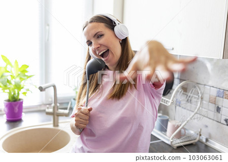 Woman in earphones listening to music and singing while cooking in the kitchen Woman in earphones listening to music and singing while cooking in the kitchen 103063061