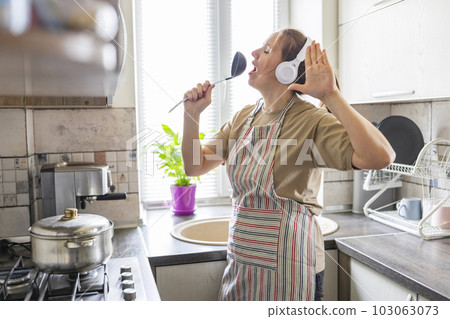 Woman in earphones listening to music and singing while cooking in the kitchen Woman in earphones listening to music and singing while cooking in the kitchen 103063073